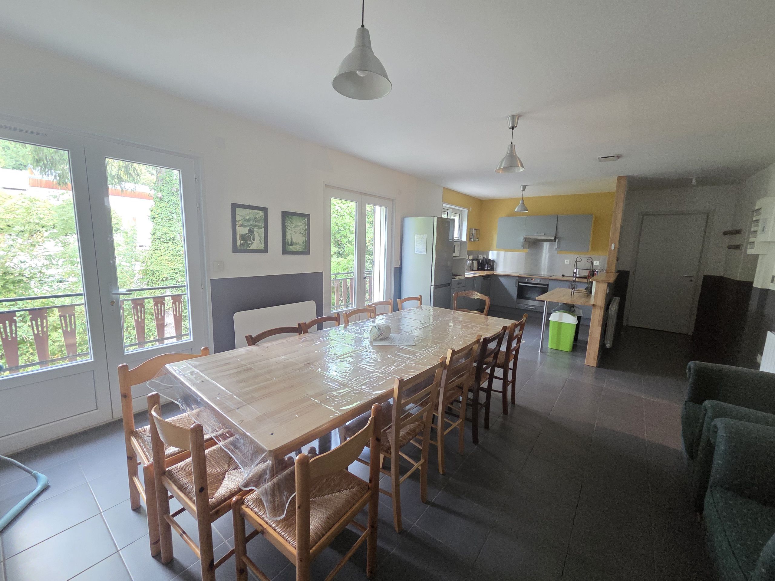 Un vaste espace de salle à manger avec une grande table et des chaises, une cuisine à l’arrière-plan et un balcon offrant une vue sur la verdure.