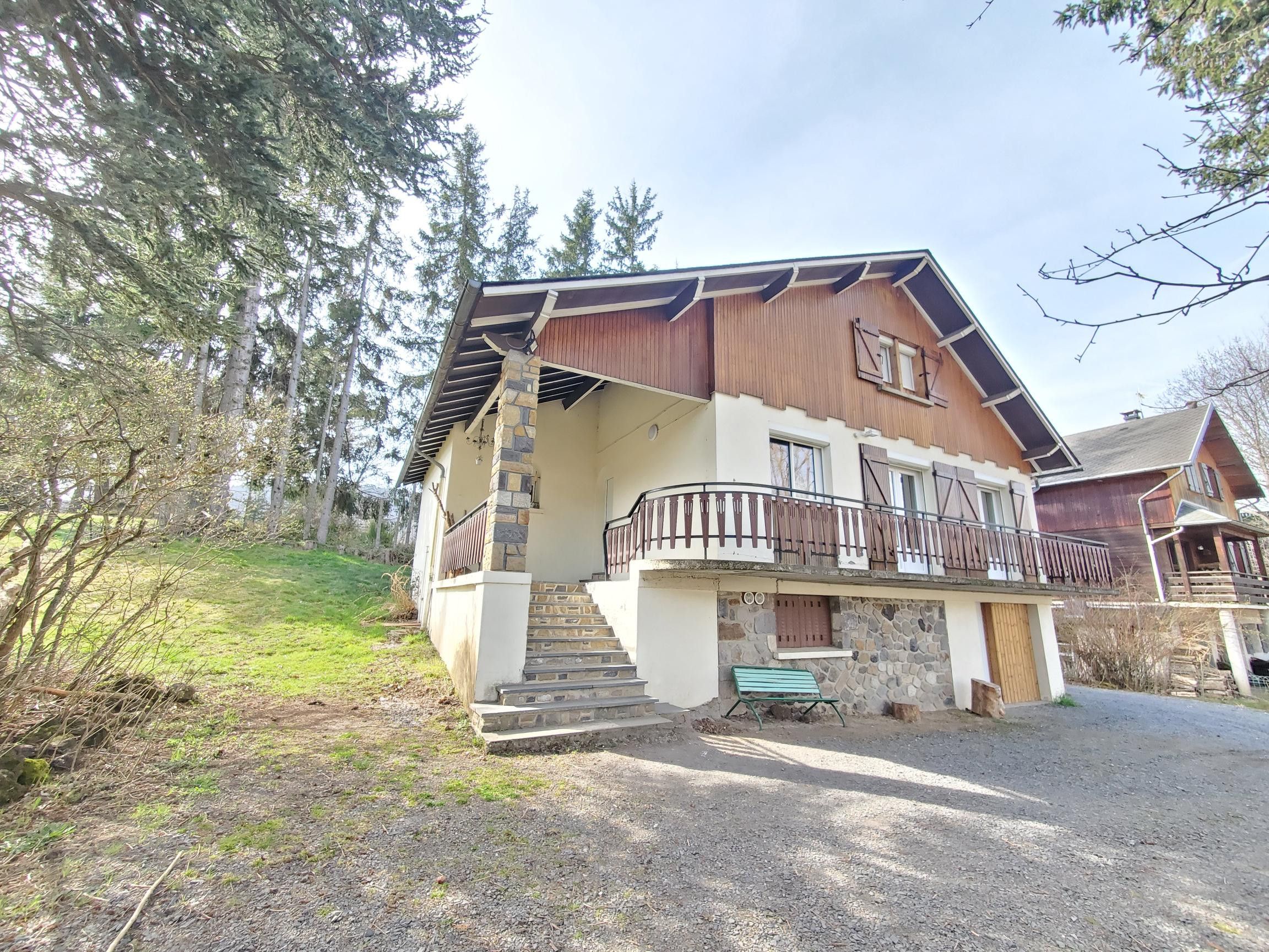 A two-story chalet with a stone staircase leading to a wooden deck, surrounded by trees.