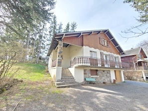 A two-story chalet with a stone staircase leading to a wooden deck, surrounded by trees.