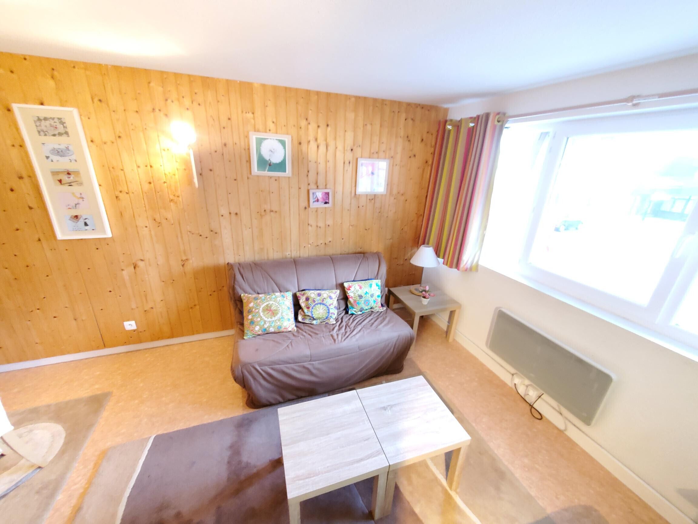 A cozy room with a wooden wall, a bed with patterned pillows, a small table, and a window with curtains.