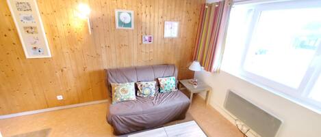A cozy room with a wooden wall, a bed with patterned pillows, a small table, and a window with curtains.