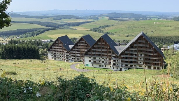 A row of wooden chalets with balconies, set in a lush green valley with distant hills.