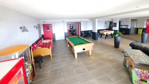 A room with a pool table, a foosball table, and a billiards table. There are chairs and a sofa.
