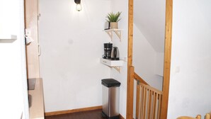 Habitació petita i neta amb terra de fusta, paret blanca, una petita estanteria amb una planta, una cafetera i un cendrer.