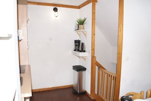 A small, clean room with a wooden floor, a white wall, a small shelf with a plant, a coffee maker, and a trash bin. - 4149a (Besse-et-Saint-Anastaise)