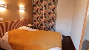 Habitació d'hotel amb llit, capçalera de fusta, dues làmpades penjades a la paret, cortina amb motius florals i una porta blanca.
