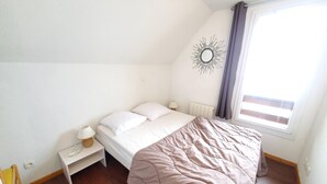 A bedroom with a bed, a nightstand, a lamp, a window with curtains, and a wall decoration. - 4145a (Besse-et-Saint-Anastaise)