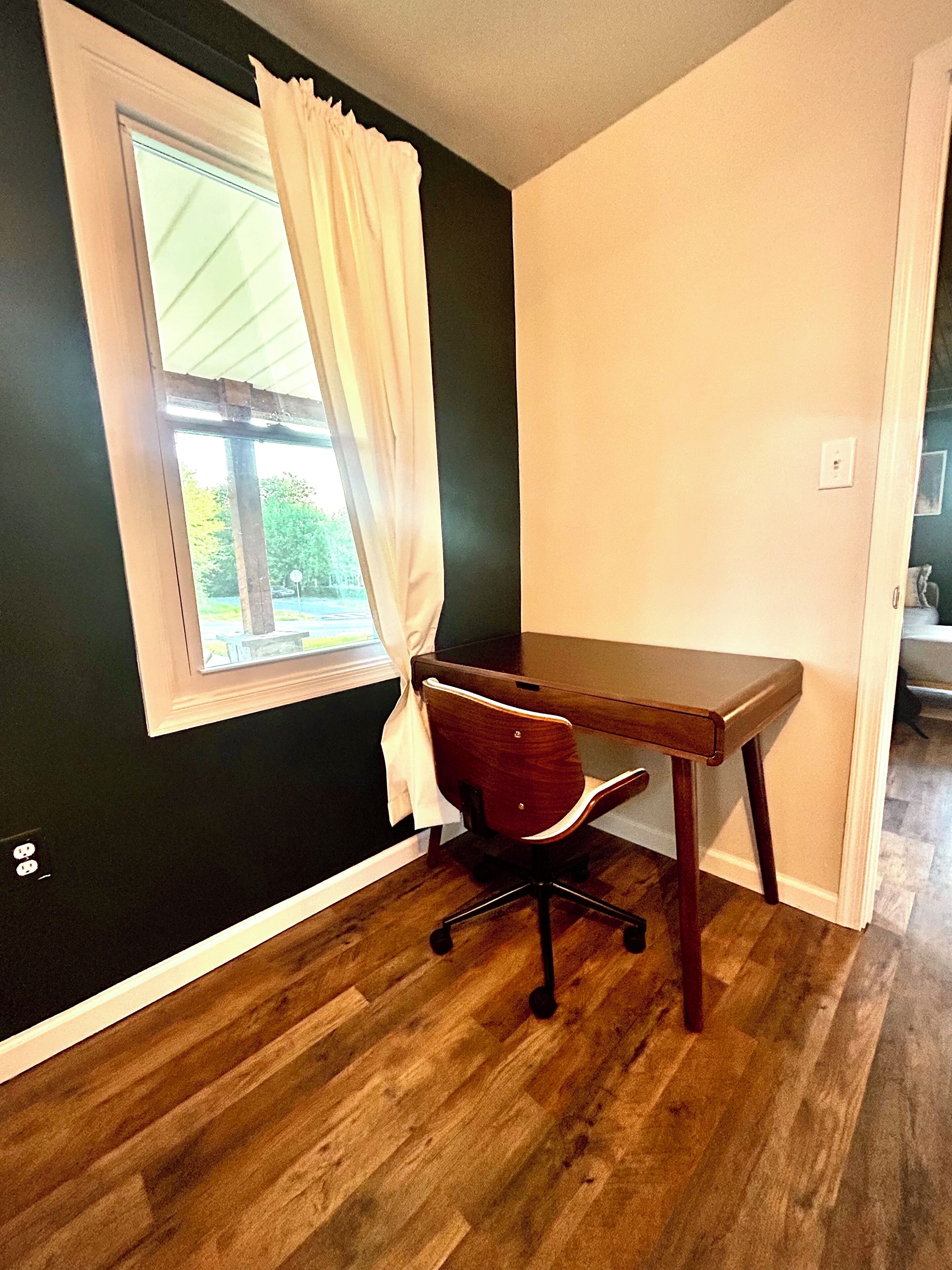 Quarto com escrivaninha de madeira, uma cadeira, janela com cortinas brancas e vista para o verde lá fora.
