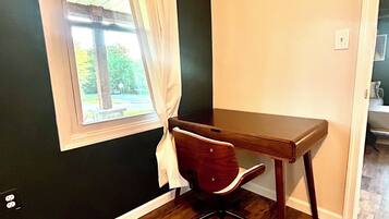 Quarto com escrivaninha de madeira, uma cadeira, janela com cortinas brancas e vista para o verde lá fora.