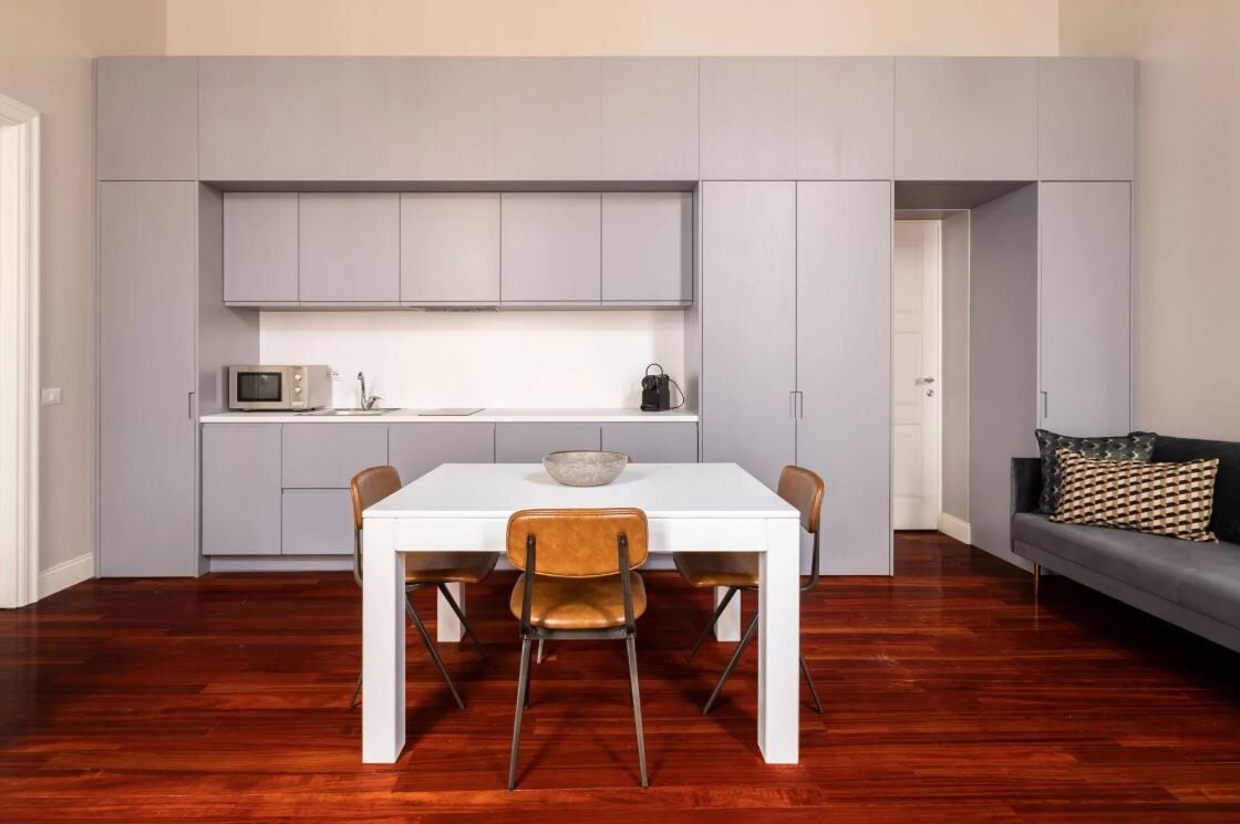 Cocina pequeña con armarios grises, encimera blanca y un microondas. Hay una mesa de comedor con dos sillas y un bol encima. Un sofá gris con un cojín estampado se ve al fondo.