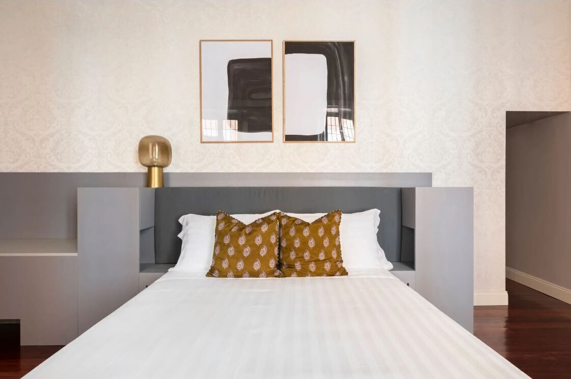 A modern hotel room with a large bed, a grey headboard, and two framed abstract paintings above the bed.