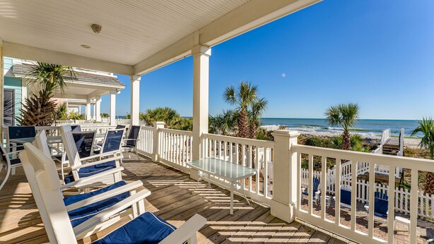 A covered outdoor seating area with white railings and blue cushions, overlooking a beach with palm trees and the ocean. - 7-Bedroom Home w/ Private Pool & Ocean Views (Surfside Beach)