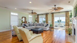 A spacious living room with a sectional sofa, a chaise lounge, and a fireplace. There are two ceiling fans, large windows with curtains, and a view of the ocean through the sliding glass doors. - 7-Bedroom Home w/ Private Pool & Ocean Views (Surfside Beach)
