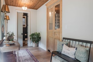Um quarto com teto de madeira, um banco com almofadas, um abajur, uma mesa com uma tigela e plantas, e uma porta que dá para outro cômodo.
