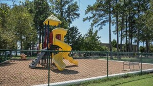 A playground with a yellow slide and climbing structure, surrounded by a fence and trees. - The Container Home at River Rocks Landing (Gadsden)