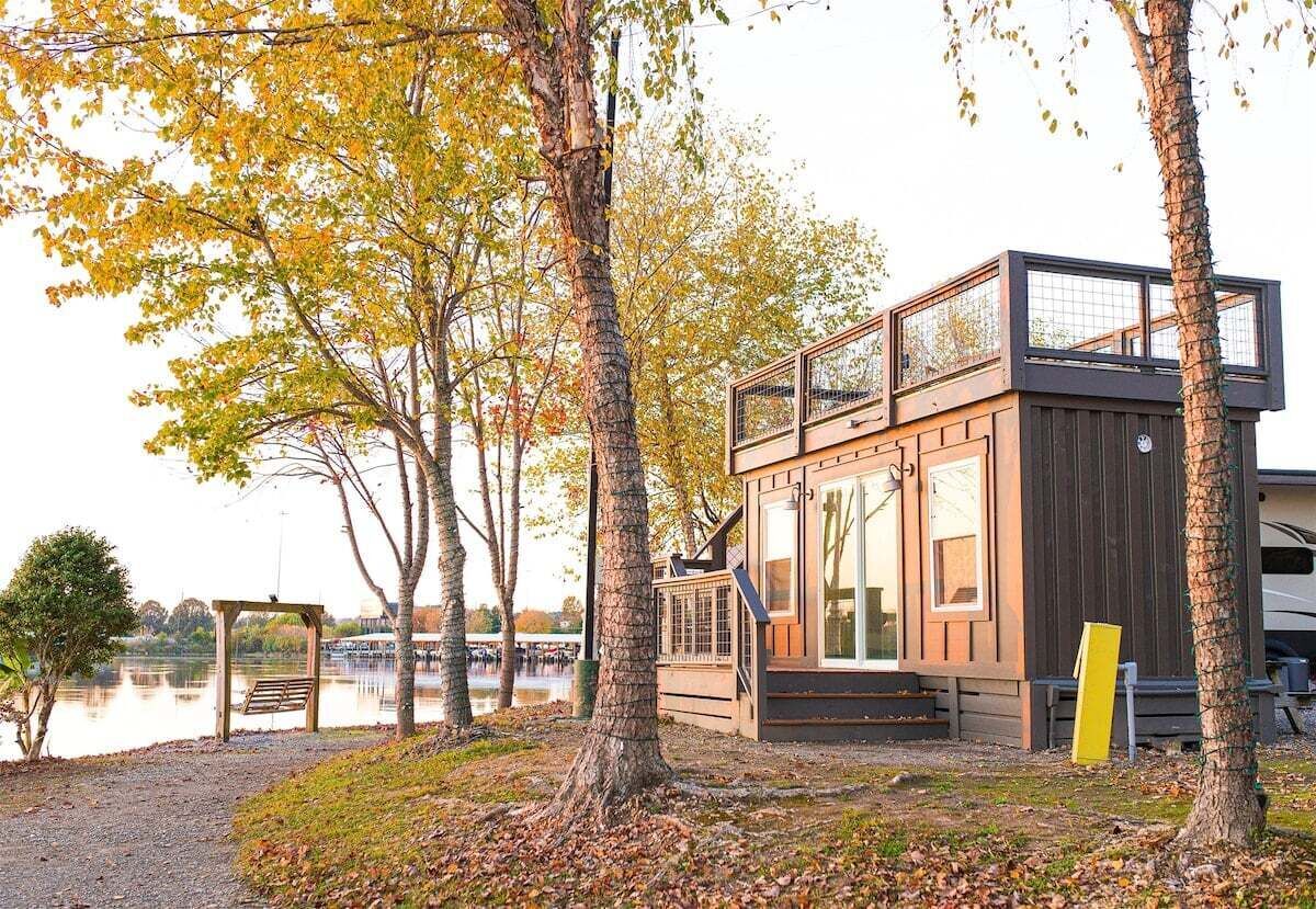 Une cabane moderne avec un étage supérieur en verre, située au bord d’un lac, entourée d’arbres et agrémentée d’un banc.