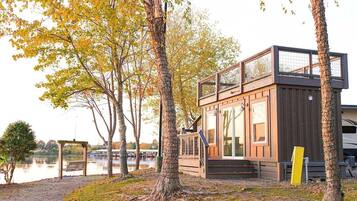 A modern cabin with a glass upper floor, situated by a lake with trees and a bench.