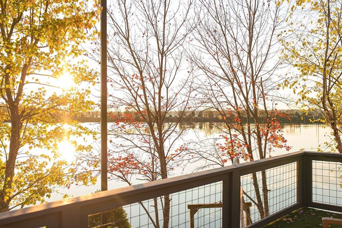 Une vue sur un lac, avec des arbres aux couleurs automnales, une terrasse en bois et une rambarde en métal.