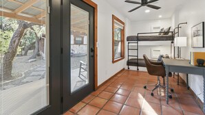 Een kleine kamer met een stapelbed, een bureau met stoel, een plafondventilator en een raam met jaloezieën.