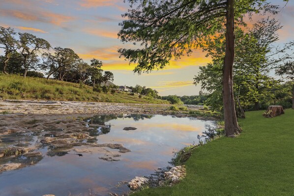 Une rivière dont les berges sont bordées de rochers et d’herbe, entourée d’arbres et d’une zone herbeuse.