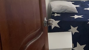 Ein Zimmer mit Holztür, einem Bett mit Sternenmuster-Überwurf, einem weißen Nachttisch und einem Fenster mit vertikalen Jalousien.