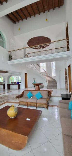 A spacious interior with a high ceiling, a staircase leading to an upper level, and a seating area with a sofa and a coffee table. - Villa Martha Garza Blanca with Chef & Full Staff (Puerto Vallarta)