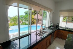 A kitchen with a large window overlooking a swimming pool, a sink, and a countertop. - Villa Martha Garza Blanca with Chef & Full Staff (Puerto Vallarta)