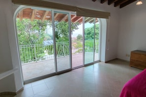 Quarto com uma grande porta de vidro deslizante que dá para uma varanda com guarda-corpo de metal e vegetação lá fora.