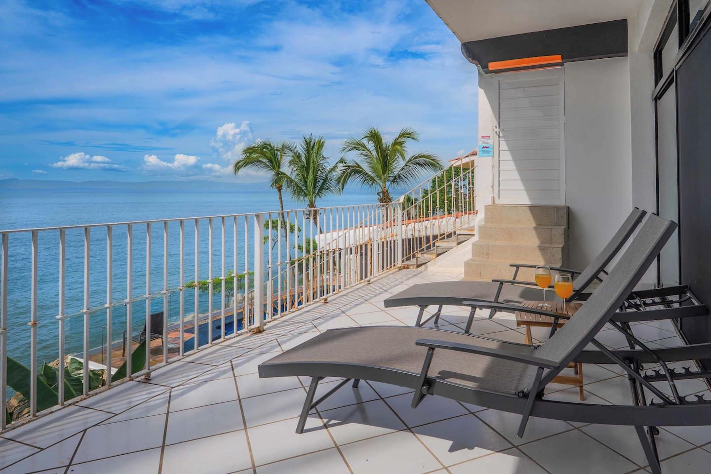 A balcony with a view of the sea, two lounge chairs, and a small table with drinks.
