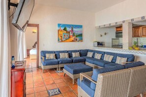 Una sala de estar con un sofá seccional grande, una zona de cocina con salpicadero de azulejos y un cuadro en la pared.
