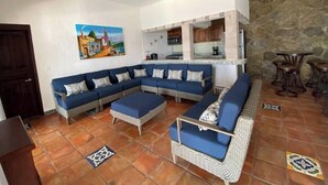A living room with a large L-shaped sofa, a small coffee table, and a kitchen area with wooden cabinets and a stone wall.