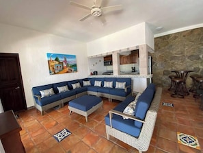 A living room with a large L-shaped sofa, a small coffee table, and a kitchen area with wooden cabinets and a stone wall.