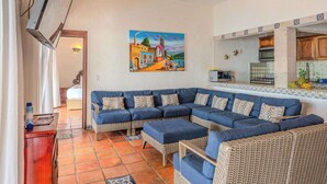 A living room with a sectional sofa, a dining area with a kitchen in the background, and a painting on the wall.