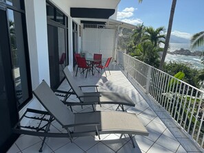 A balcony with sun loungers and a table, overlooking a coastal view with palm trees and a rocky shoreline. - Hacienda + Condo 4: Oceanfront, Sleeps 18 (Aguacate)