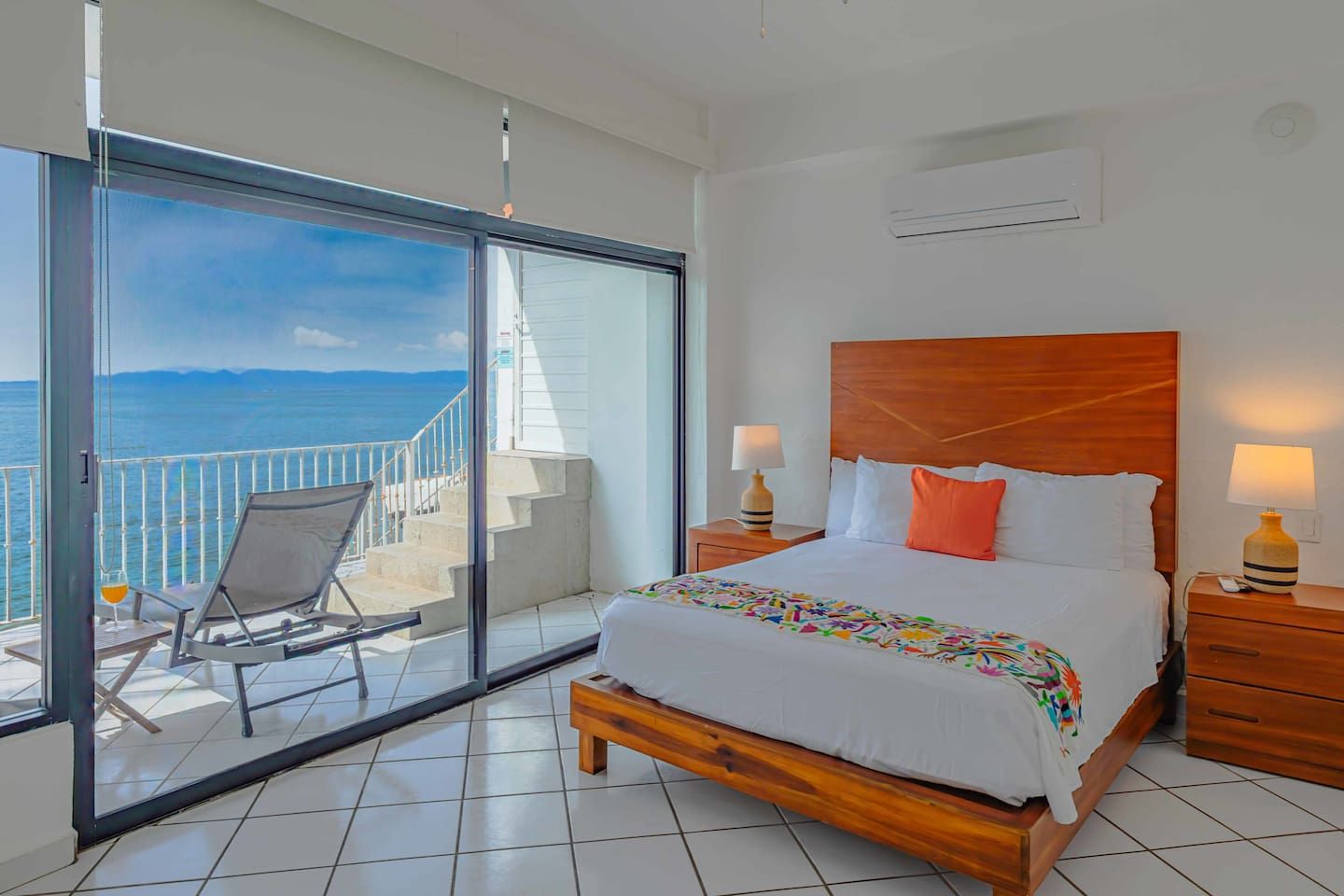 A hotel room with a bed, two bedside tables with lamps, a chair, a balcony with a view of the sea, and a large sliding glass door.