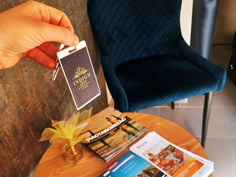 Una mano che tiene una keycard con la scritta “INDIUM CITY PANOR”, un tavolo di legno con brochure, un vaso con fiori gialli e una sedia blu scuro.