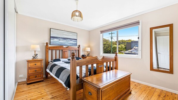 Une chambre à coucher avec un lit en bois, des tables de chevet, une commode, une coiffeuse, une lampe suspendue, un tableau encadré et une fenêtre donnant sur des bateaux et la verdure.