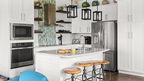 Una cocina moderna con armarios blancos, electrodomésticos de acero inoxidable, una isla central con taburetes e iluminación colgante decorativa.