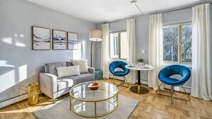 Uma sala de estar moderna com um sofá cinzento, duas poltronas azuis, uma mesa de centro de vidro redonda e uma pequena mesa redonda com uma luminária.