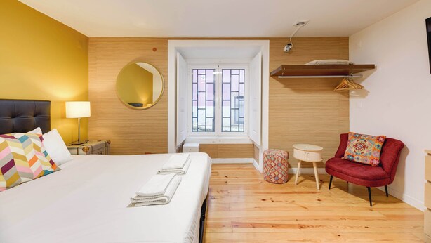 A hotel room with a bed, a bedside table with a lamp, a red chair, a small round table, and a wooden floor.