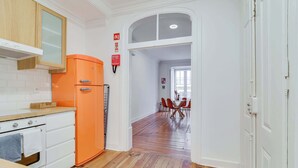 Une cuisine avec des meubles blancs, un réfrigérateur orange, un plancher en bois et un coin repas comprenant une table et des chaises.