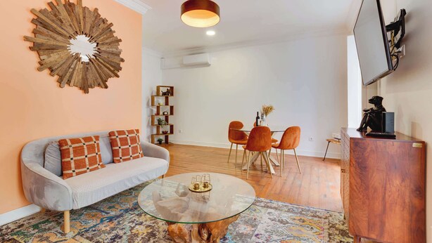 Un salon moderne avec une table basse ronde en verre, un canapé gris agrémenté de coussins à motifs, une décoration murale en bois, un coin repas avec des chaises orange et un téléviseur à écran plat fixé au mur.