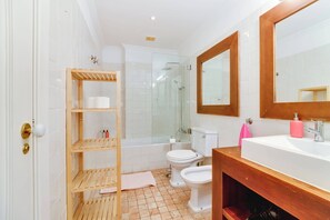 Un baño con piso de baldosas blancas, estante de madera con artículos de tocador, inodoro blanco, ducha con mampara de vidrio y tocador de madera con espejo.