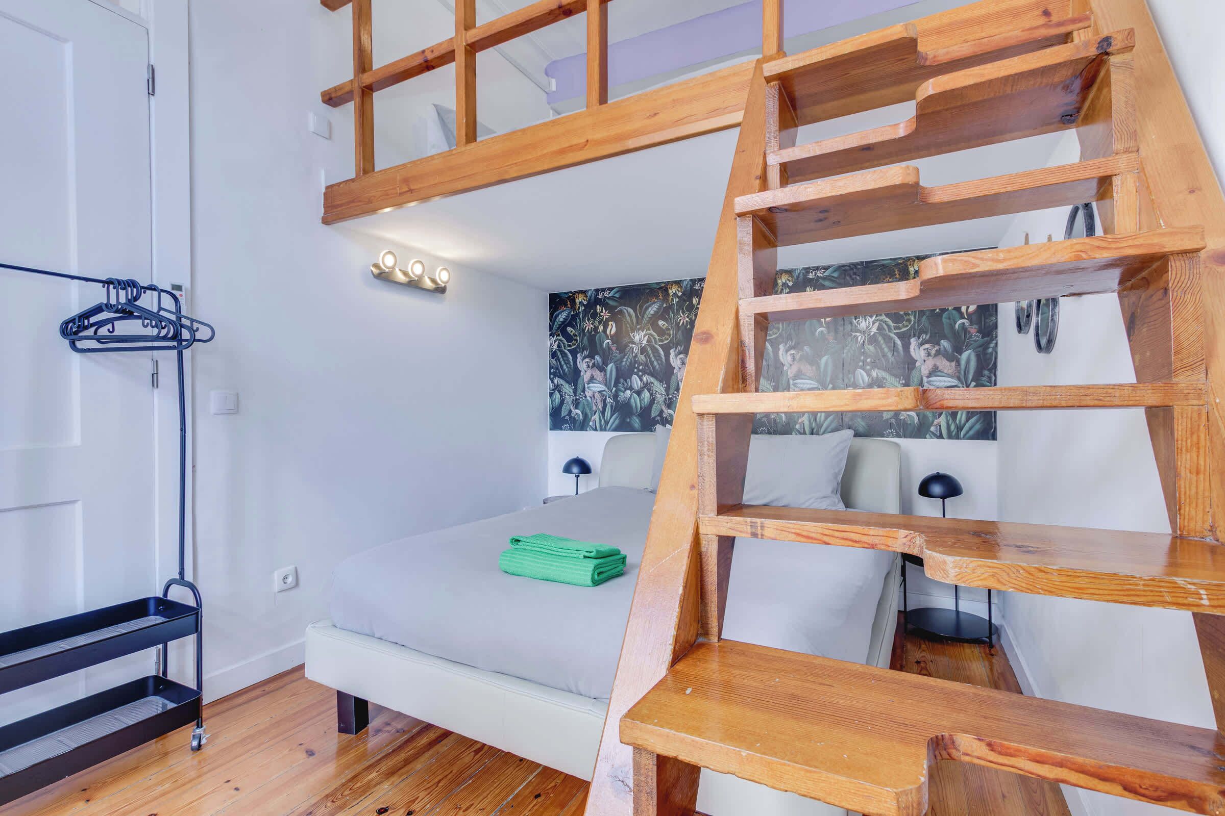 Un dormitorio con una escalera de madera, una cama con una toalla verde, una lámpara de pared y un perchero negro.