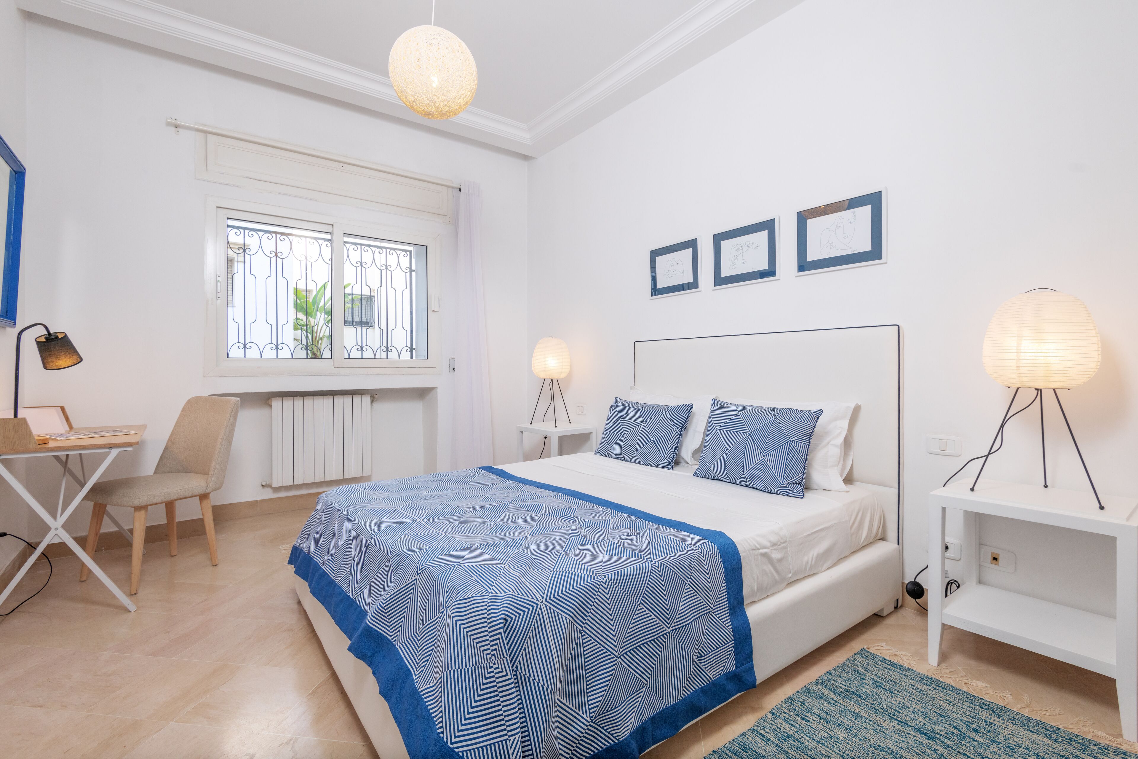 A bedroom with a bed, a desk, and a chair. There are two bedside lamps and a window with metal bars.