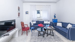 A modern living room with a large flat-screen TV, a blue sofa, a red chair, and a coffee table with decorative items. - L'Oiseau Bleu Marsa Plage  (La Marsa)