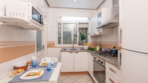 A compact kitchen with white cabinets, a stove, and a sink. There is a table with plates and glasses, and a window with a view of the outside. - L'Oiseau Bleu Marsa Plage  (La Marsa)