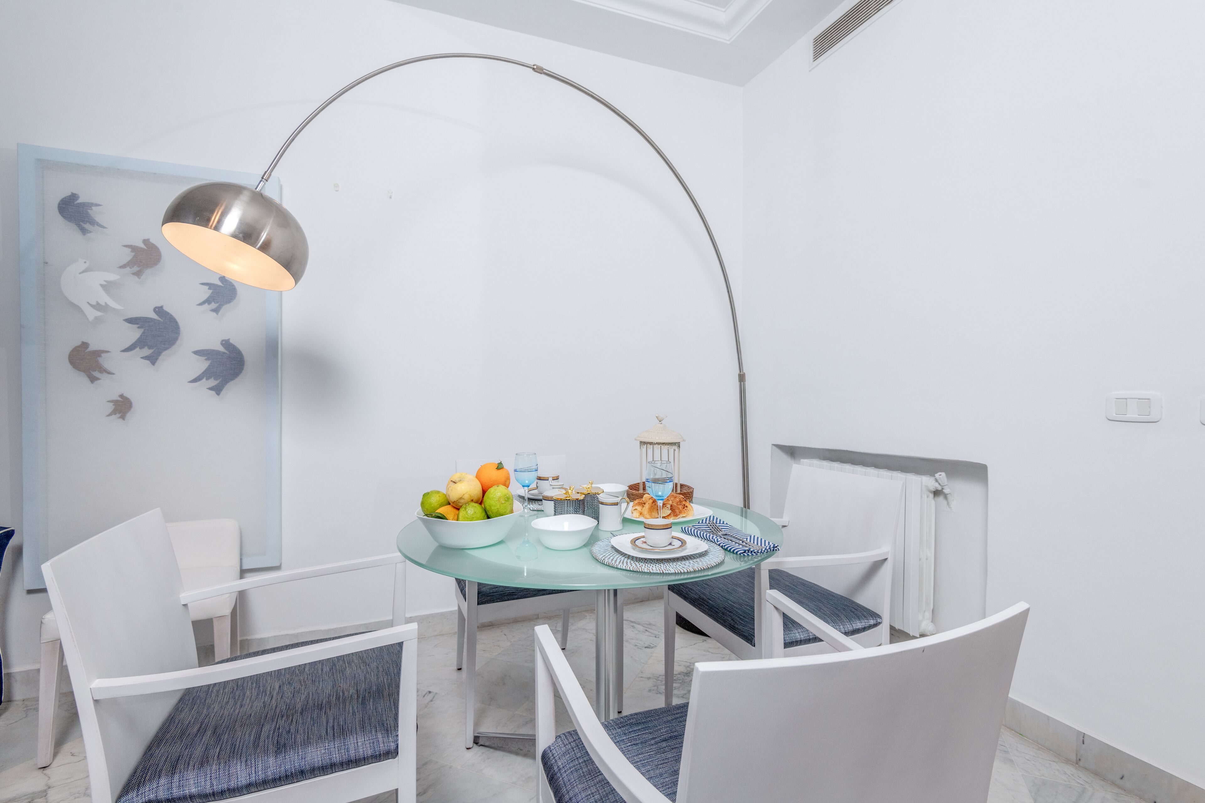 A dining area with a round glass table, white chairs with blue cushions, a metal lamp, and a wall with shark silhouettes.