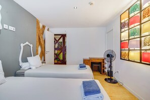 Habitació d'hotel amb dos llits, una porta de fusta, escriptori, ventilador i una paret amb làmpades de disseny.