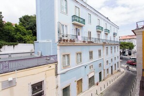 A multi-story building with balconies, a street in front, and a tree-lined background.